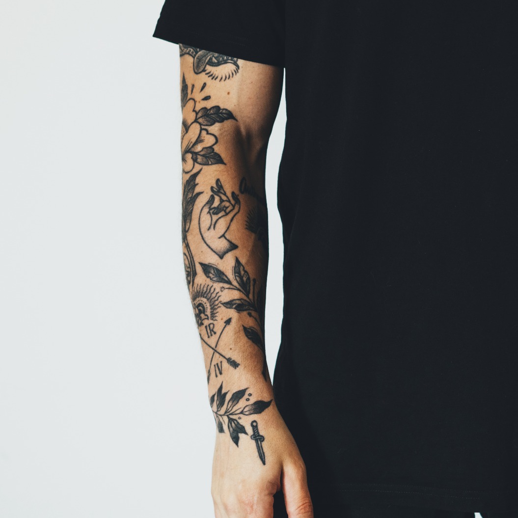 Ein junger, attraktiver, bärtiger Mann mit Tattoos, bekleidet mit einem schwarzen, leeren T-Shirt, posiert vor einem weißen Wandhintergrund. Leerer Raum für dein Logo oder Design.